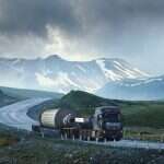 Large white transport truck transporting commercial cargo in semi trailer running on turning way highway road with scenic mountains mountaineous scenery in background.